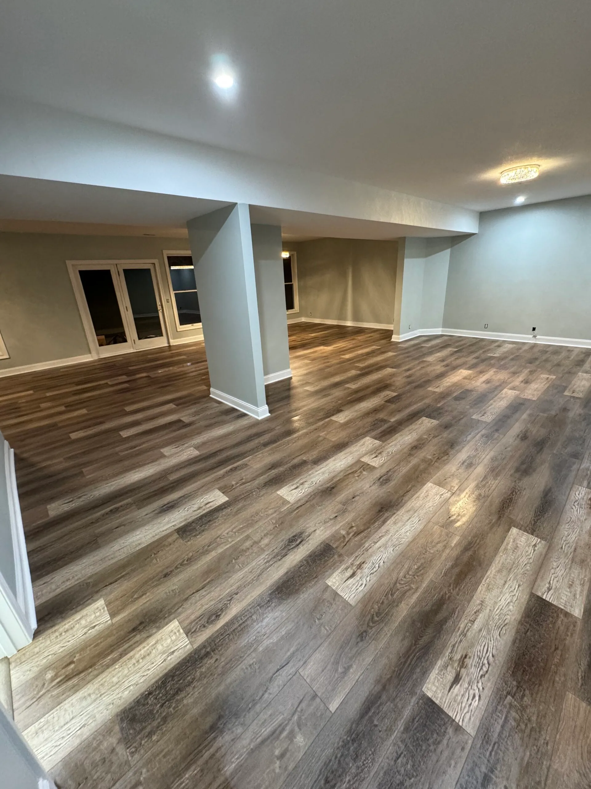 Weathered-oak look luxury vinyl plank running continuously through a large finished basement with support columns and egress windows