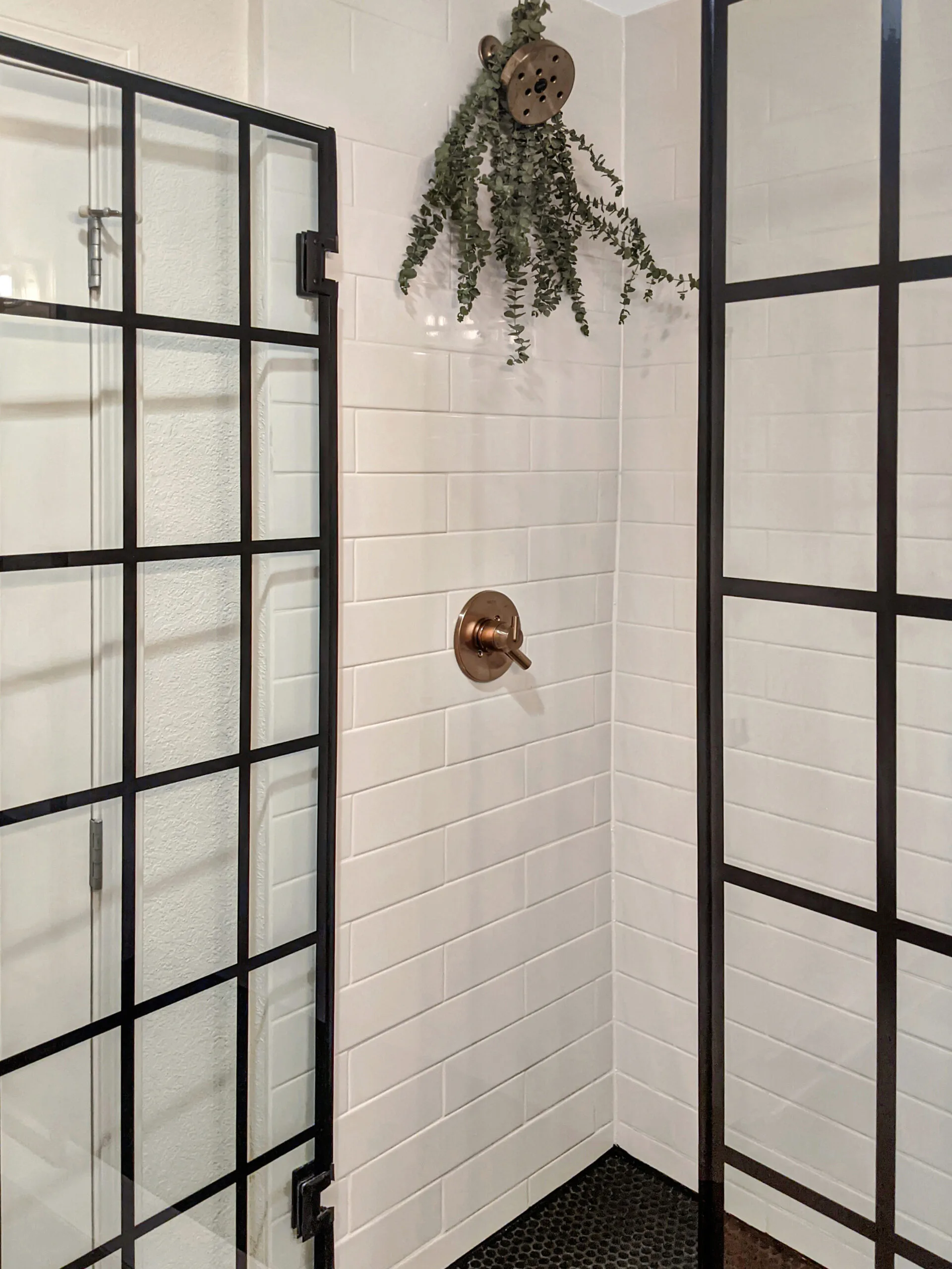 White subway tile shower laid in a running bond pattern with an aged brass shower head and valve, seen through a black-framed glass door