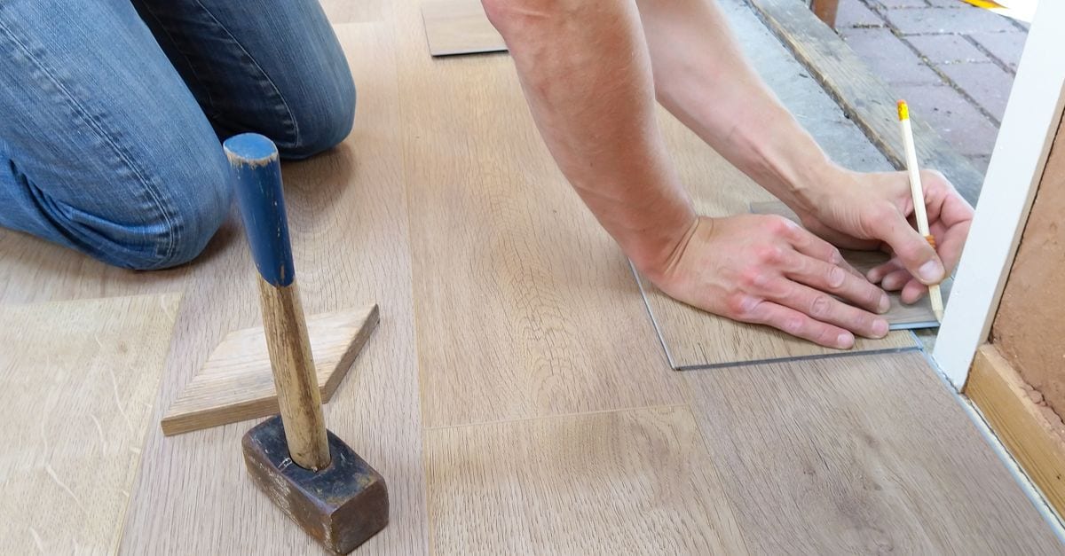 Installer kneeling to measure and mark a light oak plank with a pencil at a doorway, with a tapping block and mallet resting on the finished floor beside them