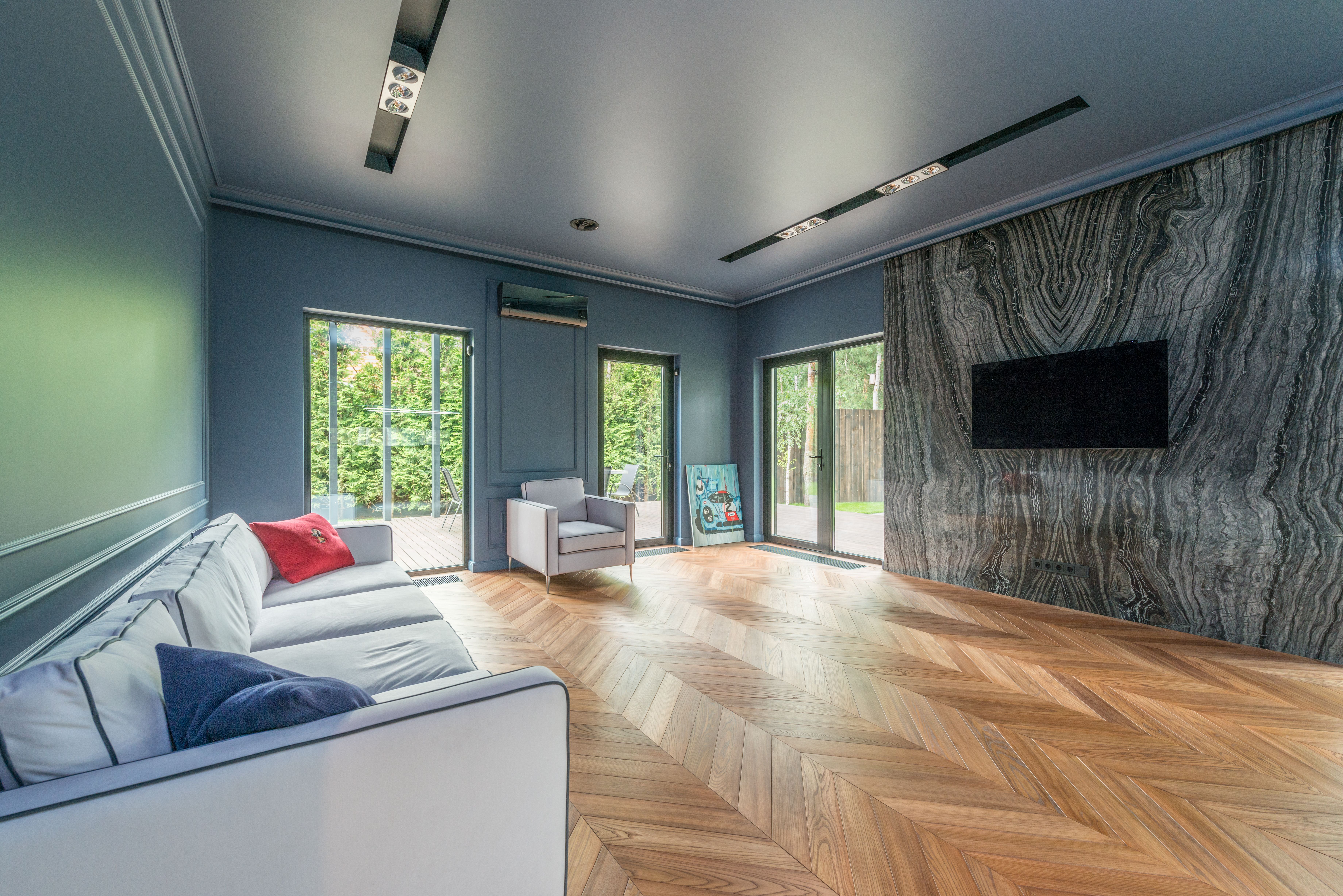 Light oak floor laid in a chevron pattern running through a living room with blue-grey panelled walls, a pale sofa, and glass doors onto a garden