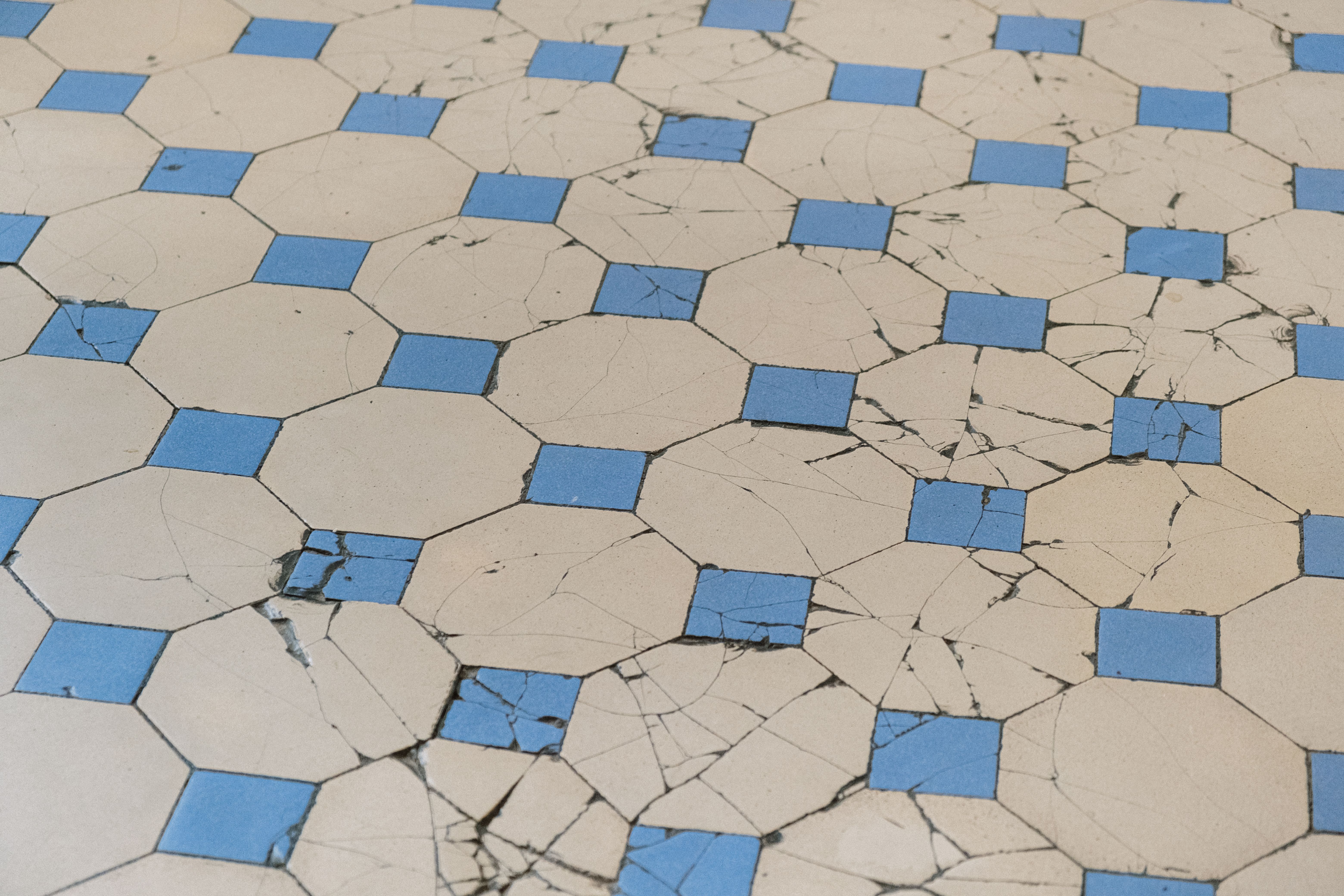 Beige octagon-and-blue-dot ceramic floor tile with long fractures running through tiles and grout broken away at several joint intersections
