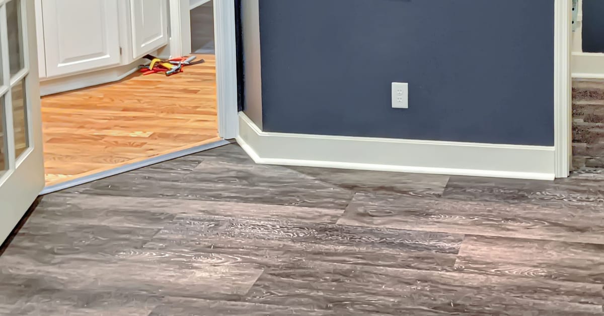 Gray luxury vinyl plank meeting an existing red oak floor at a doorway, joined by a low metal transition strip, with new white baseboard along a navy wall