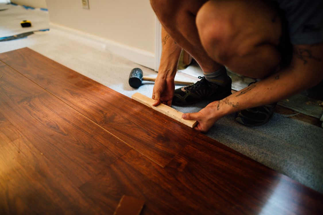 Worker kneeling to tap a dark wood plank into place with a tapping block and rubber mallet over grey underlayment